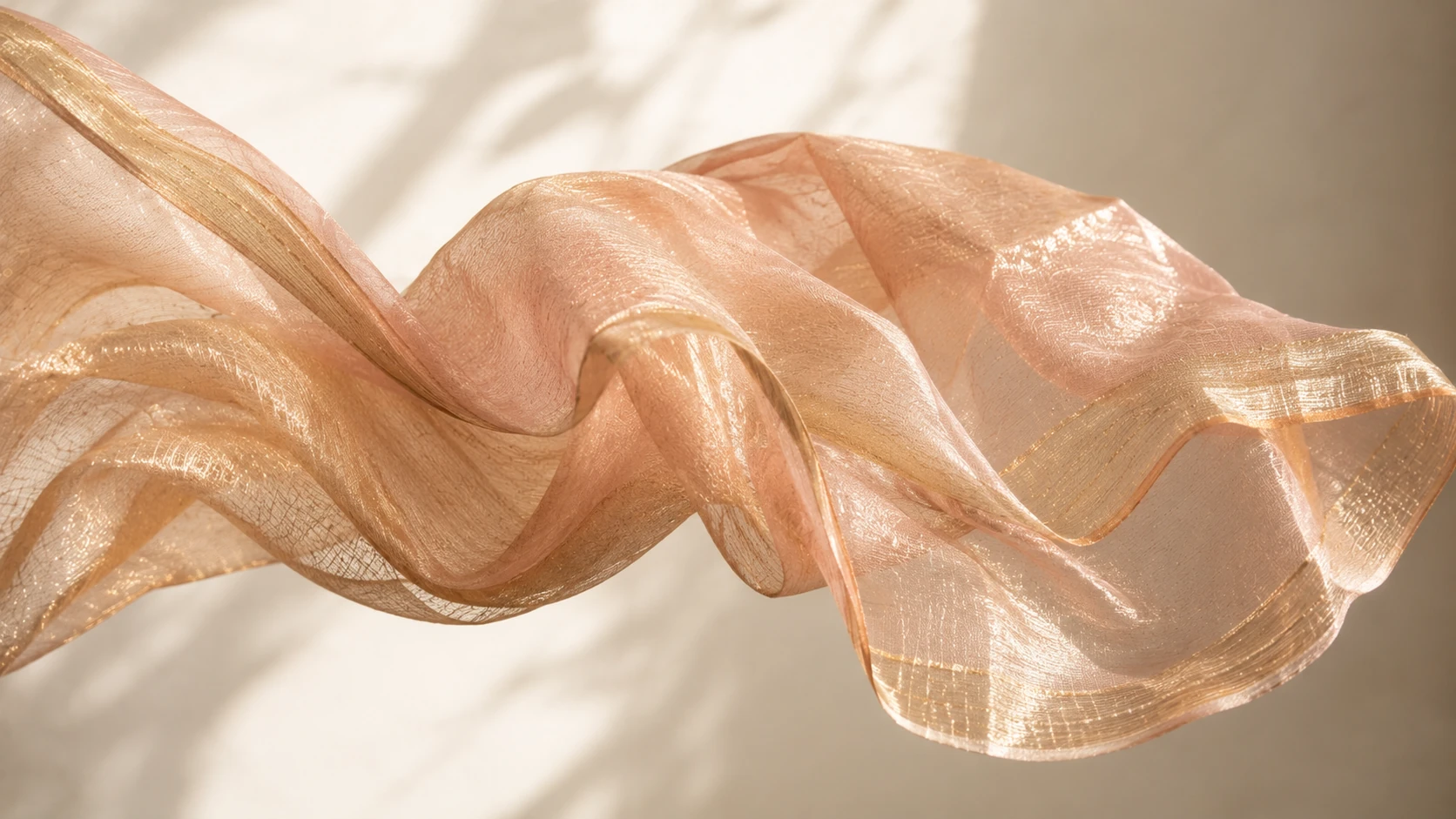 Blush and gold tissue fabric draped mid-air showing its metallic shimmer and sheer drape